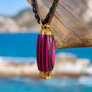 Hearthwood urn pendant with pink camo laminate wood and gold stainless steel core hanging from a black braided necklace on a weathered wooden post by bright blue ocean water