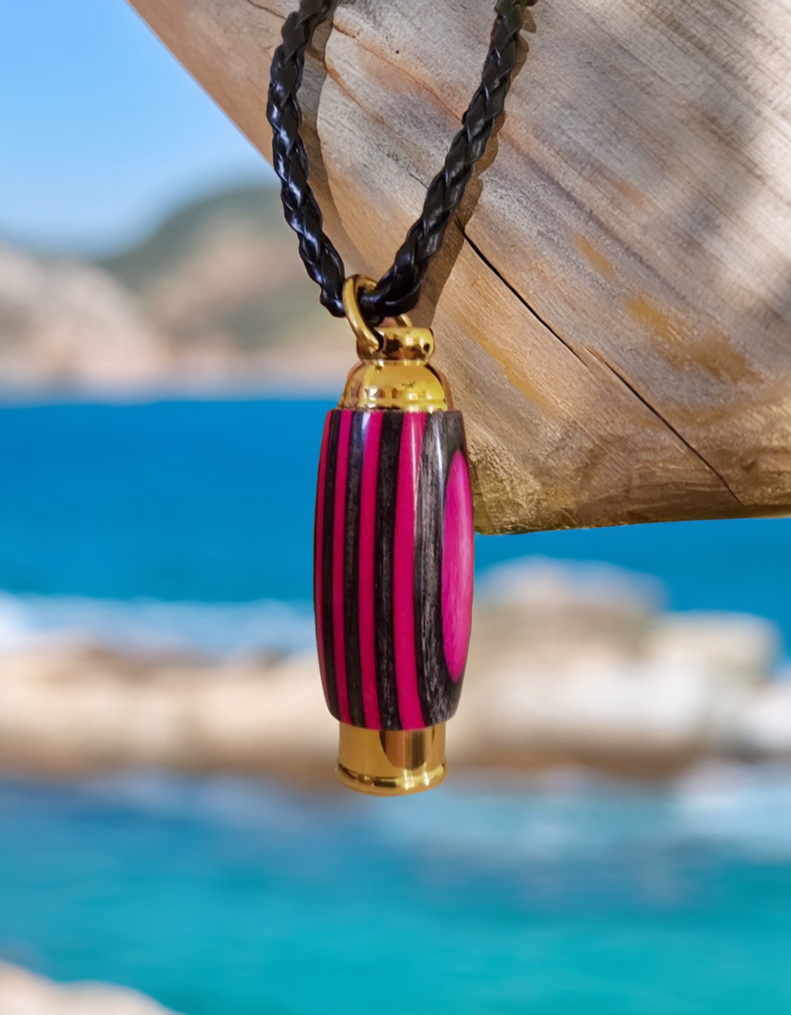 Hearthwood urn pendant with pink camo laminate wood and gold stainless steel core hanging from a black braided necklace on a weathered wooden post by bright blue ocean water