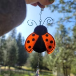 Hand holding a ladybug suncatcher with bright orange wings and black spots, featuring infused cremation ash and a small crystal drop, photographed outdoors in sunlight.