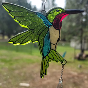 Hummingbird suncatcher with bright green glass wings, blue body, cream chest, red throat detail, and a dangling crystal prism, shown hanging outdoors in natural daylight