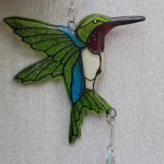 Hummingbird suncatcher with green glass wings, blue and white body, red throat detail, and dangling crystal prism, hanging against a light wood background