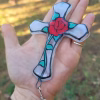 Handheld stained-glass cross suncatcher featuring a red rose design with cremation ash infused, resting in an open palm with a crystal drop hanging below.