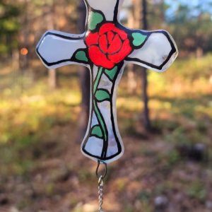 Red rose cross suncatcher handcrafted with cremation ash, featuring a painted red rose and green leaf design on translucent white glass with a crystal drop accent, shown outdoors in natural light.