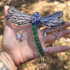Dragonfly suncatcher with green glass body, hand-painted wings, and dangling crystal prisms, displayed resting in an open hand outdoors in natural sunlight