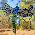Dragonfly suncatcher with green glass body, blue head, hand-painted transparent wings, and multiple dangling crystal prisms, hanging outdoors against a sunlit woodland background