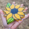 Sunflower suncatcher handcrafted with cremation ash, featuring bright yellow petals, green leaves, and crystal teardrop accents displayed in an open hand.