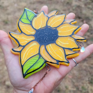 Sunflower suncatcher handcrafted with cremation ash, featuring bright yellow petals, green leaves, and crystal teardrop accents displayed in an open hand.