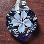 Handcrafted deep blue glass cremation pendant featuring a detailed white flower suspended above swirling white flecks of encased cremation ashes, shown in a close-up view to highlight texture and color