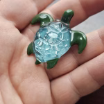 Handcrafted glass sea turtle cremation pendant with green flippers and an aqua shell containing white flecks of encased cremation ashes, shown resting in the hand to highlight size and intricate detail