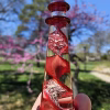 A hand holding a red glass lighthouse infused with cremation ash, featuring a spiraling red core with shimmering white ash suspended throughout. The lighthouse is photographed outdoors against a bright blue sky, with blooming pink trees and soft greenery blurred in the background.