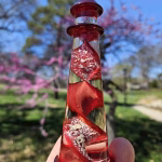 A hand holding a red glass lighthouse infused with cremation ash, featuring a spiraling red core with shimmering white ash suspended throughout. The lighthouse is photographed outdoors against a bright blue sky, with blooming pink trees and soft greenery blurred in the background.