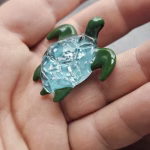 Handcrafted glass sea turtle cremation pendant with green flippers and an aqua shell containing white flecks of encased cremation ashes, shown resting in the hand to highlight size and intricate detail