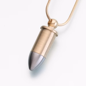 Close-up of a bullet-shaped cremation pendant featuring a brushed brass-toned body and a smooth stainless steel tip, hanging from a gold snake chain against a light background.