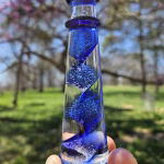 A hand holding a cobalt-blue glass lighthouse infused with cremation ash, featuring a spiral core of swirling blue and white bubbles. The lighthouse is photographed outdoors with blooming pink trees and bright spring greenery softly blurred in the background.