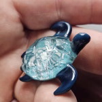 Handcrafted glass sea turtle cremation pendant with deep blue fins and an aqua shell filled with white flecks of encased cremation ashes, shown resting in the hand to highlight size and detail