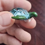 Handcrafted glass sea turtle cremation pendant shown from the side, with a green head and flippers and an aqua shell containing white flecks of encased cremation ashes, held between fingers for scale