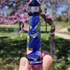 A hand holding a cobalt-blue glass lighthouse infused with cremation ash, featuring a spiral core of swirling blue and white bubbles. The lighthouse is photographed outdoors with blooming pink trees and bright spring greenery softly blurred in the background.