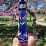 A hand holding a cobalt-blue glass lighthouse infused with cremation ash, featuring a spiral core of swirling blue and white bubbles. The lighthouse is photographed outdoors with blooming pink trees and bright spring greenery softly blurred in the background.
