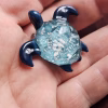 Hand-crafted glass sea turtle cremation pendant with dark blue fins and an aqua shell containing white flecks of encased cremation ashes, shown resting in the palm for scale