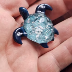 Hand-crafted glass sea turtle cremation pendant with dark blue fins and an aqua shell containing white flecks of encased cremation ashes, shown resting in the palm for scale