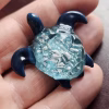Hand-crafted glass sea turtle cremation pendant with dark blue fins and an aqua shell containing white flecks of encased cremation ashes, shown resting in the palm for scale