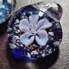 Handcrafted glass flower cremation pendant featuring a delicate white blossom suspended above a deep blue background with white flecks of encased cremation ashes, shown in a close-up to highlight detail and color