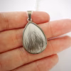 Pear-shaped memorial pendant filled with preserved hair or fur, shown resting in the palm of a hand with a silver chain in the background.