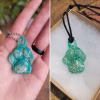 Teal glass paw print cremation pendant with visible encased cremation ash shown on a silver ball chain in a hand and on a black cord inside a small gift box