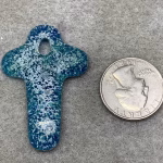 Glass cross cremation pendant in Cyan and Royal Blue glass with encased cremation ash, shown beside a quarter for size reference on gray felt background