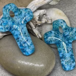 Glass cross cremation pendants in cyan and royal blue glass with encased cremation ash, displayed on smooth river stone with driftwood background