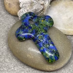 Glass cross cremation pendant in blue and green glass with encased cremation ash, displayed on smooth river stone with driftwood background