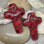 Glass cross cremation pendants in deep ruby red glass with encased cremation ash, displayed on smooth river stones with driftwood background