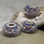 Glass cremation beads in deep purple, blue, and multicolor tones with visible encased ash and sterling silver grommets, arranged on driftwood and smooth stone
