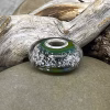 Glass cremation bead in layered green and charcoal tones with visible encased ash and a sterling silver grommet, displayed on smooth stone with driftwood in the background