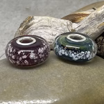 Glass cremation beads in burgundy and deep teal with visible encased ash and sterling silver grommets, displayed side by side on smooth stone with driftwood in the background