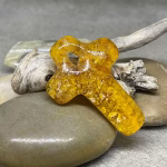 Glass cross cremation pendant in golden amber glass with encased cremation ash, displayed on smooth river stones with driftwood in the background