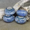 Glass cremation beads in layered blue and purple tones with visible encased ash and sterling silver grommets, arranged on driftwood against a stone background