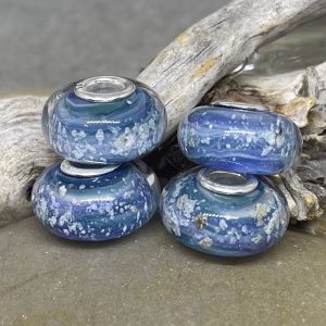 Glass cremation beads in layered blue and purple tones with visible encased ash and sterling silver grommets, arranged on driftwood against a stone background
