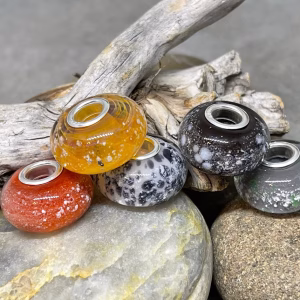 Glass cremation beads in orange, amber yellow, black, grey, and speckled white with visible encased ash and sterling silver grommets, arranged together on smooth river stones with driftwood in the background