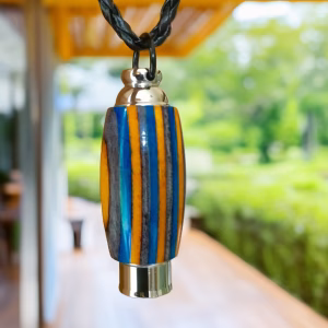 Hearthwood urn pendant with aqua fire laminate wood stripes and stainless steel core hanging from a black braided necklace outdoors with greenery and a wooden walkway in the background