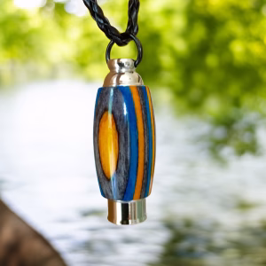 Hearthwood urn pendant with aqua fire laminate wood stripes and stainless steel core hanging from a black braided necklace outdoors near a flowing river with green foliage in the background