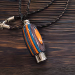 Hearthwood urn pendant with aqua fire laminate wood grain and stainless steel core resting on a dark wooden surface with black braided cord and silver chain extender, angled flat lay view
