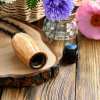 Hearthwood urn pendant with natural oak wood grain and black stainless steel core shown open with removable threaded cap resting beside it on a wood slice and rustic wooden surface with flowers in the background, flat lay view