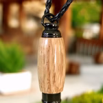 Hearthwood urn pendant with natural oak wood grain and black stainless steel core hanging from a black braided cord indoors with greenery and warm wood tones in the background, front-facing view
