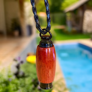 Hearthwood urn pendant in bloodwood with black stainless steel core hanging outdoors near a swimming pool on a braided black cord