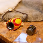 Opened Hearthwood urn cremation pendant showing the interior chamber with yellow and red layered wood body and separate black stainless steel threaded core resting on a stone surface
