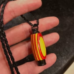 Hearthwood urn cremation pendant made with yellow and red layered wood with a black stainless steel core held in hand on a braided black cord necklace