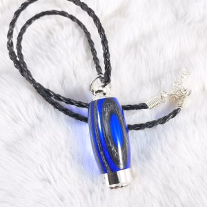 Hearthwood urn pendant with royal blue and charcoal laminate wood grain resting on a white fur background with black braided cord and silver chain extender, flat lay view