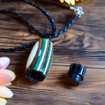 Hearthwood urn pendant with evergreen camo laminate wood grain shown open with removable threaded cap beside it on a wooden surface with black braided cord necklace and flowers, flat lay view