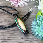 Hearthwood urn pendant with evergreen camo laminate wood grain lying on a wooden surface with black braided cord necklace and pastel flowers surrounding it, flat lay view